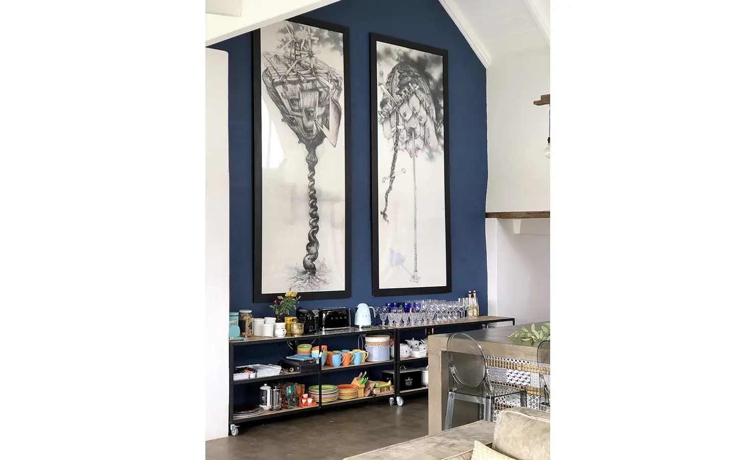 Perfect Hideaways: A navy feature wall showcases surrealist monochrome artworks above a well-stocked open shelving unit with colourful crockery and glassware, adding quirky character to the space. 22 Degrees East, Prince Albert.