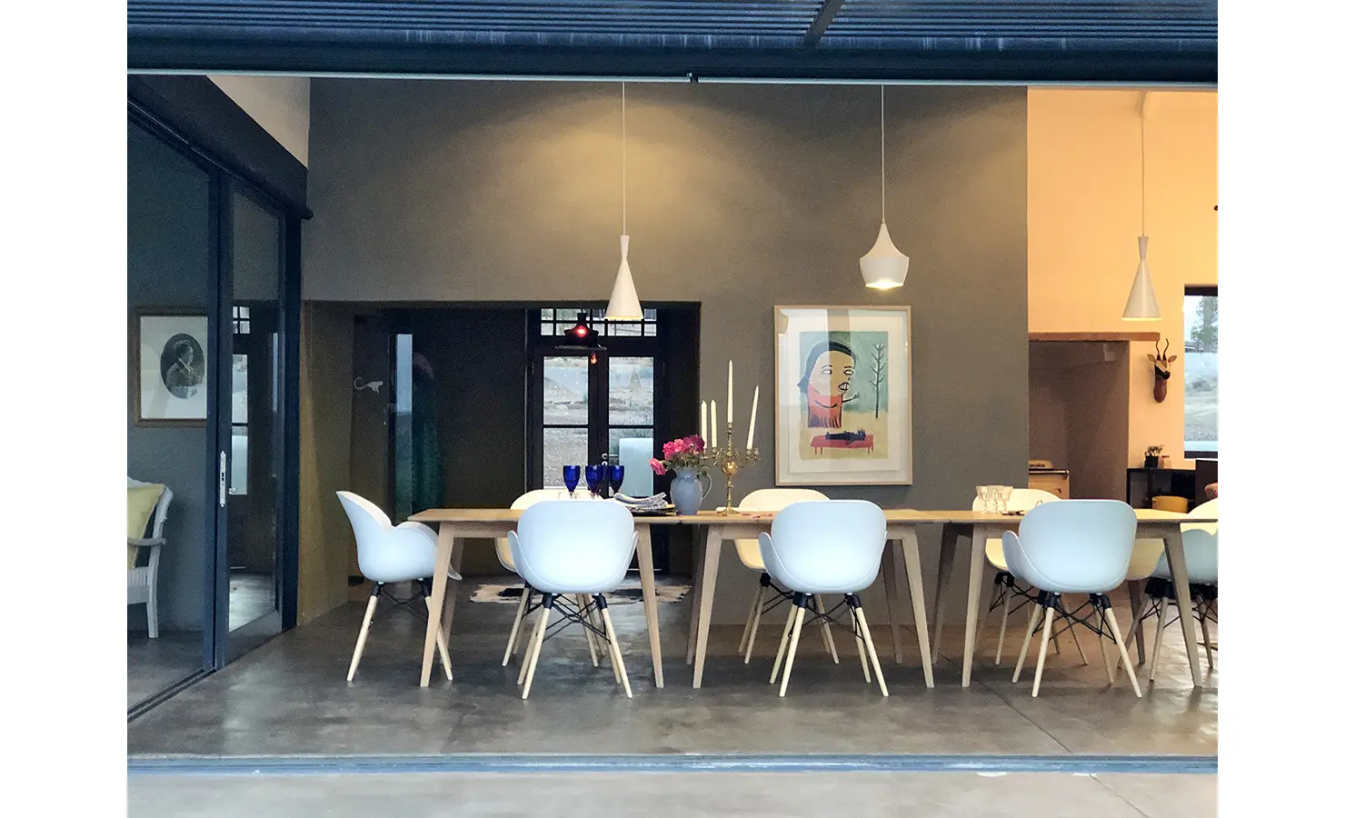 Perfect Hideaways: A modern dining room with sculptural white chairs and pale wooden table sits beneath pendant lights, framed by contemporary art and dark feature walls. 22 Degrees East, Prince Albert.