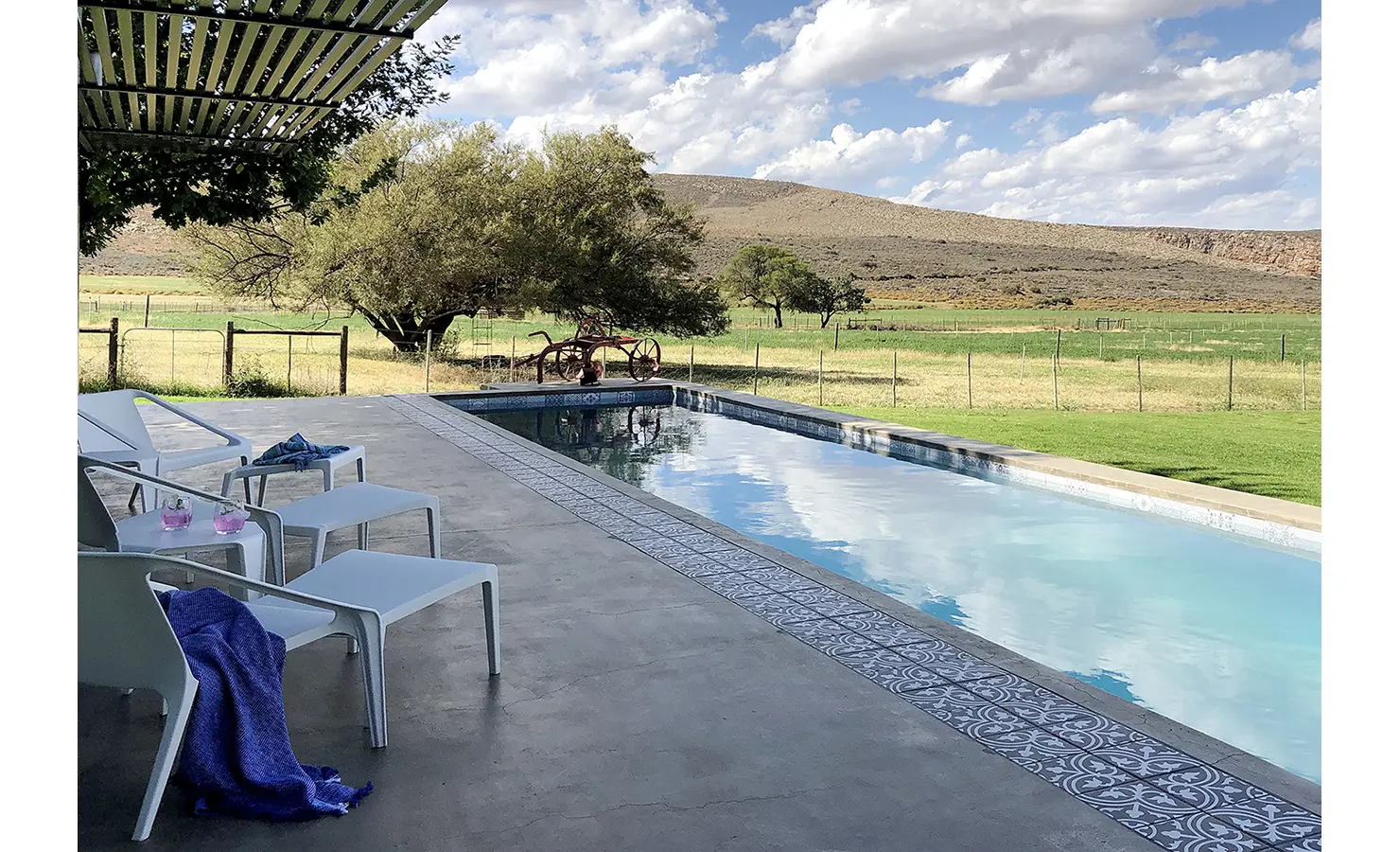 Perfect Hideaways: Poolside loungers and side tables create an inviting scene on a tiled patio overlooking green Karoo fields and distant hills under a dynamic sky. 22 Degrees East, Prince Albert.