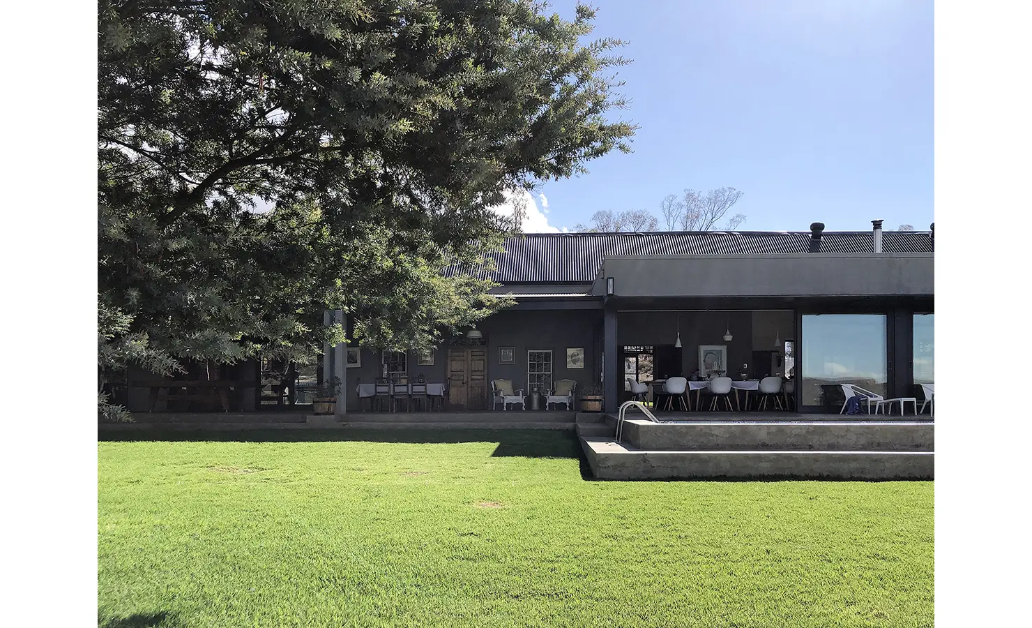 Perfect Hideaways: A modern Karoo farmhouse with a steel roof and shaded patio opens to a large green lawn and a shimmering pool, offering seamless indoor-outdoor living. 22 Degrees East, Prince Albert.