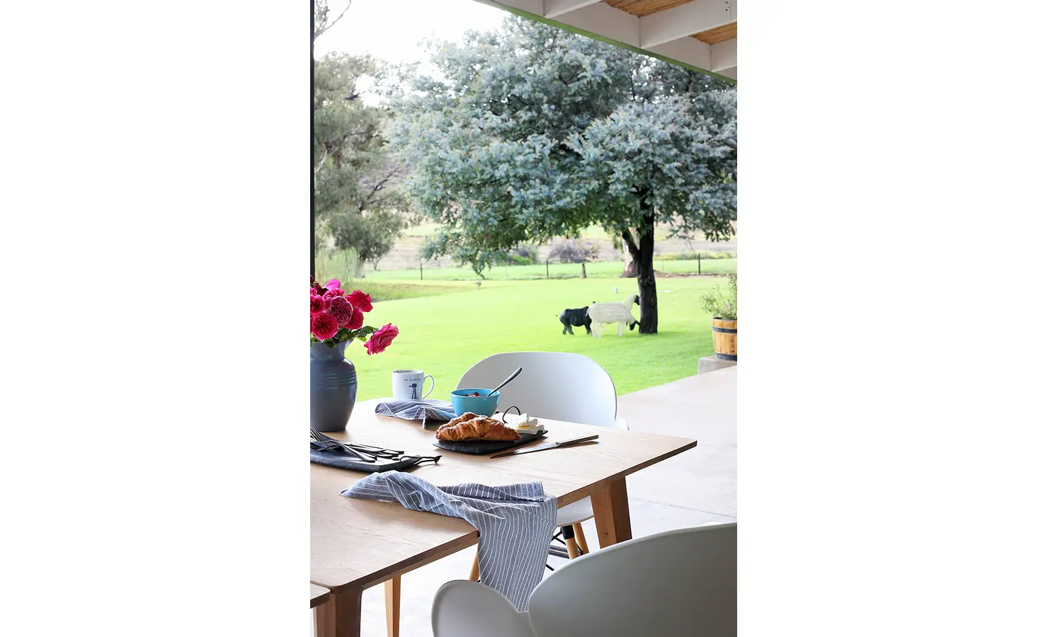Perfect Hideaways: A peaceful breakfast scene on a covered terrace features fresh croissants, bright pink roses, and soft morning light falling over a green lawn and garden sculptures. 22 Degrees East, Prince Albert.