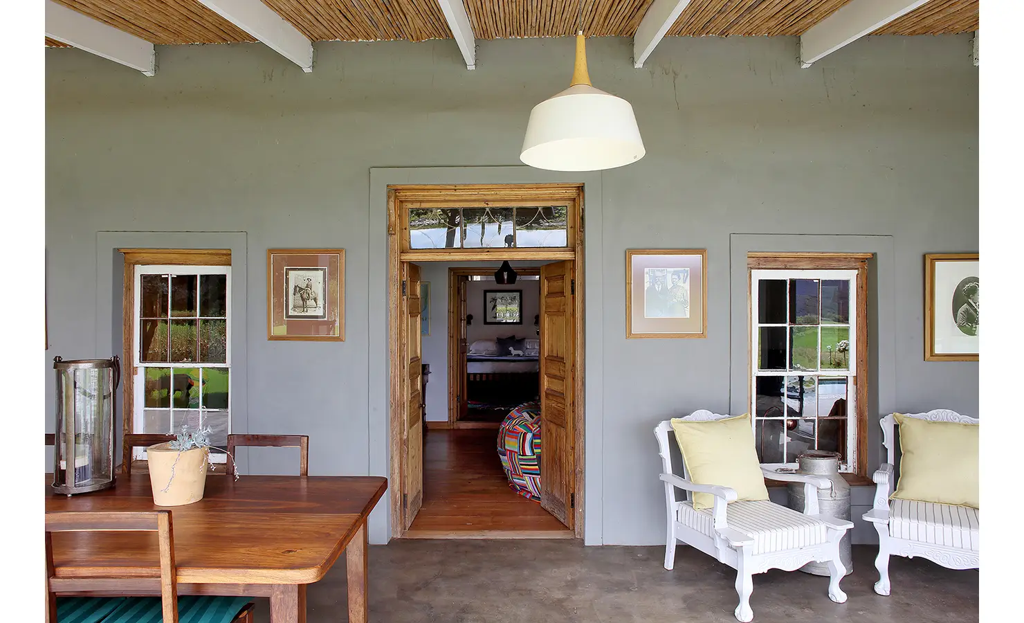 Perfect Hideaways: A rustic verandah with natural reed ceilings and framed artwork surrounds a central wooden doorway, leading into the heart of the home with a splash of colour. 22 Degrees East, Prince Albert.