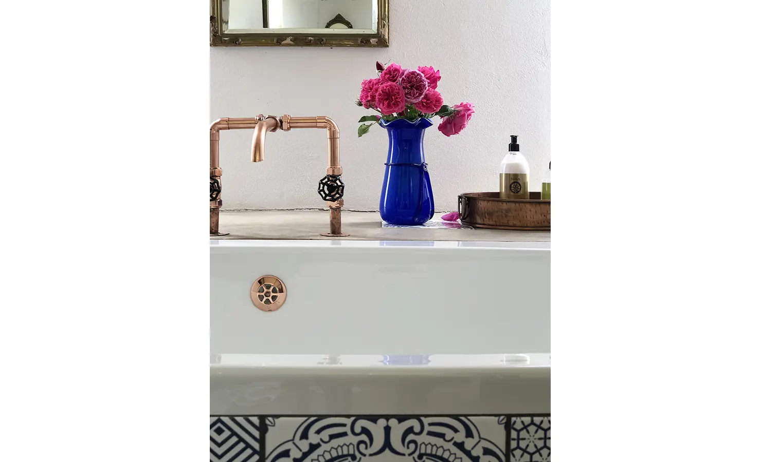 Perfect Hideaways: A deep bath with copper plumbing is set against a white wall, decorated with a royal blue vase of roses and patterned blue tiles, exuding vintage charm. 22 Degrees East, Prince Albert.