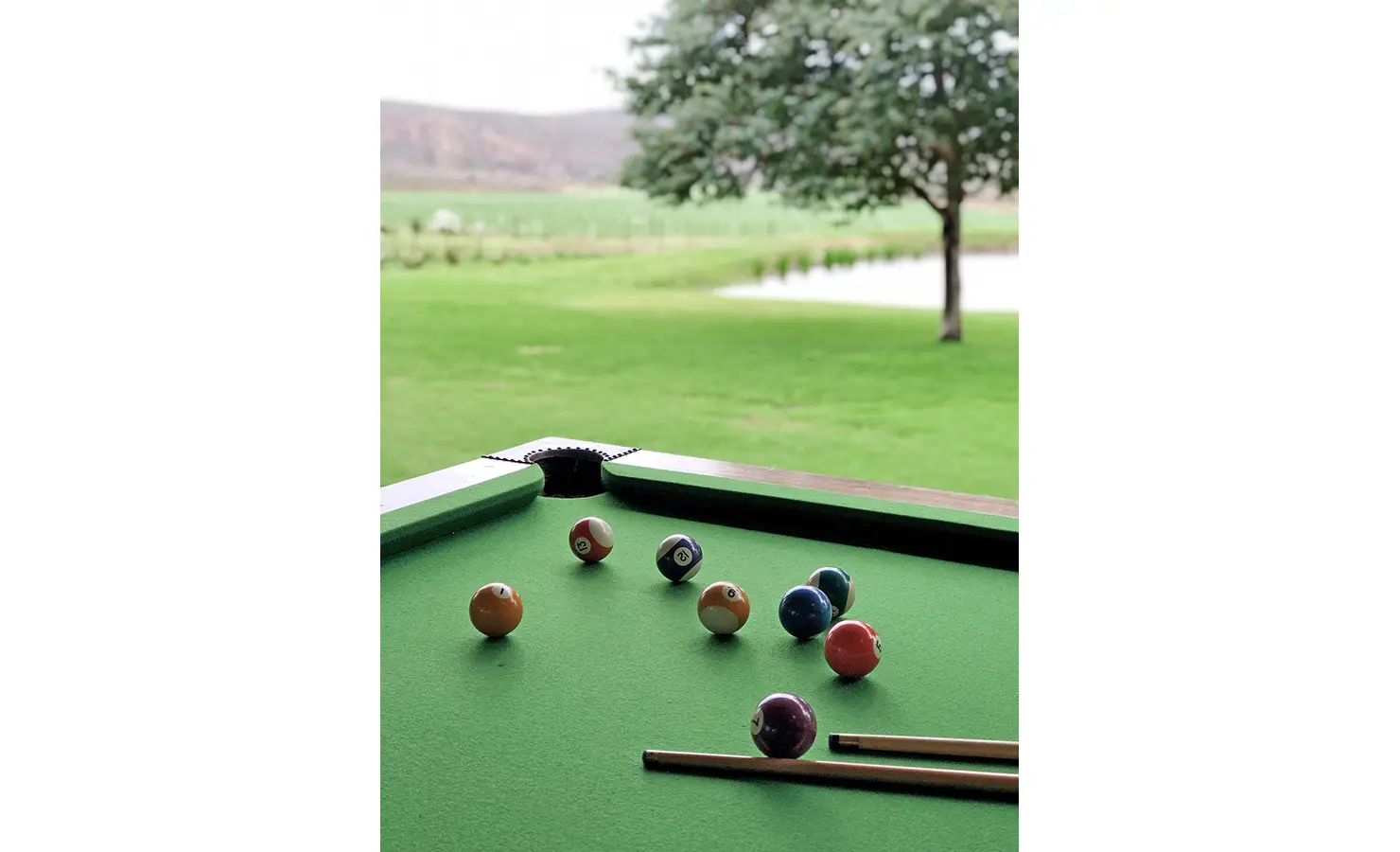 Perfect Hideaways: A pool table with vibrant green felt sits beside open doors revealing expansive views of a lush lawn, tranquil dam and surrounding mountains. 22 Degrees East, Prince Albert.