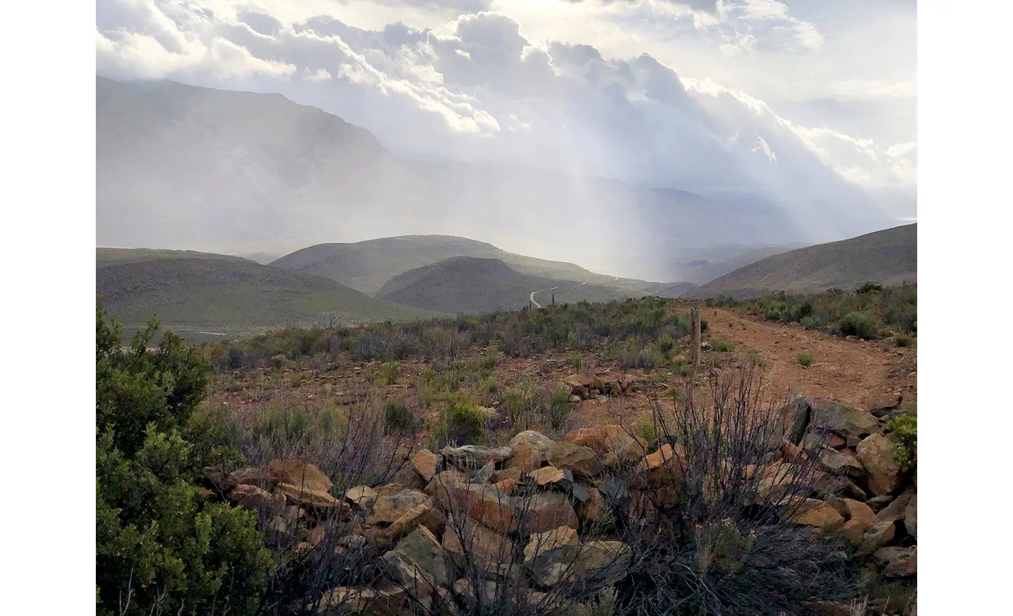 Perfect Hideaways: A misty Karoo landscape unfolds in soft light, with rays breaking through thick clouds over rolling hills and rocky terrain. 22 Degrees East, Prince Albert.