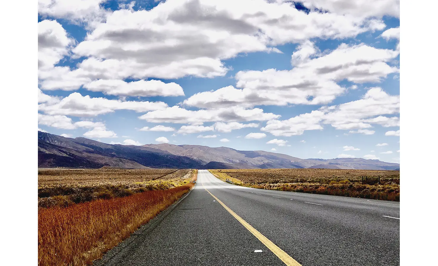 Perfect Hideaways: A long stretch of open road cuts through the vast Karoo plains, framed by golden grasses and towering mountains under a dramatic cloud-filled sky. 22 Degrees East, Prince Albert.