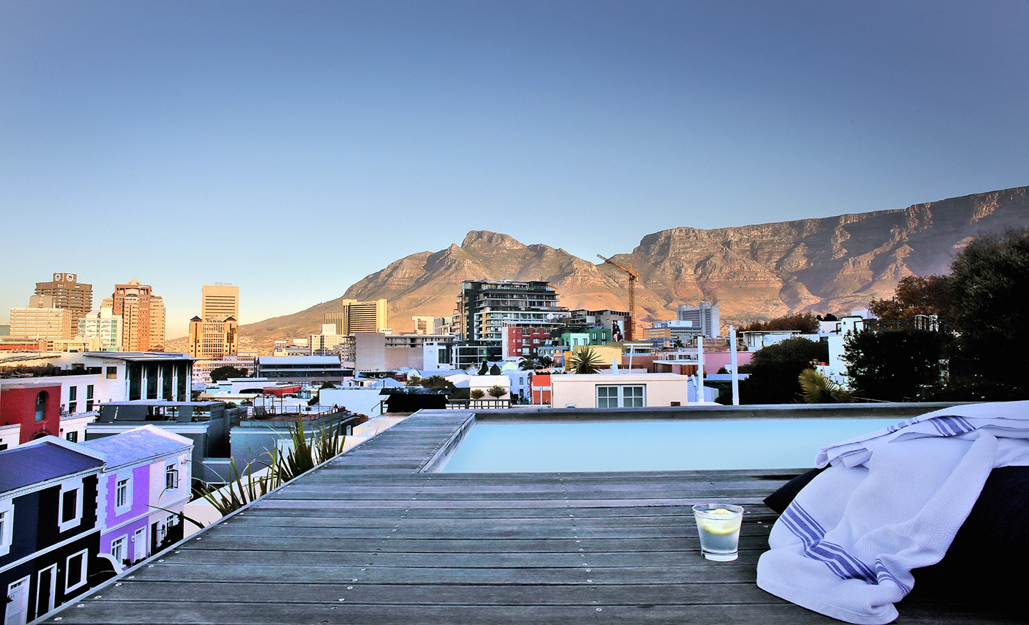 Perfect Hideaways: Rooftop deck with a plunge pool overlooking the colourful houses of De Waterkant, the Cape Town city skyline, and majestic Table Mountain bathed in late afternoon light; a towel and drink rest beside the pool, inviting relaxation. Pepper Tree House, De Waterkant.