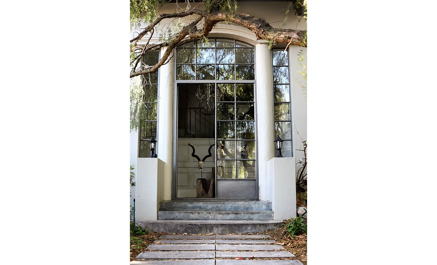 Perfect Hideaways: Grand arched entrance framed by white pillars and gridded glass panels, shaded by a pepper tree branch, creating an elegant yet natural welcome. Pepper Tree House, De Waterkant.