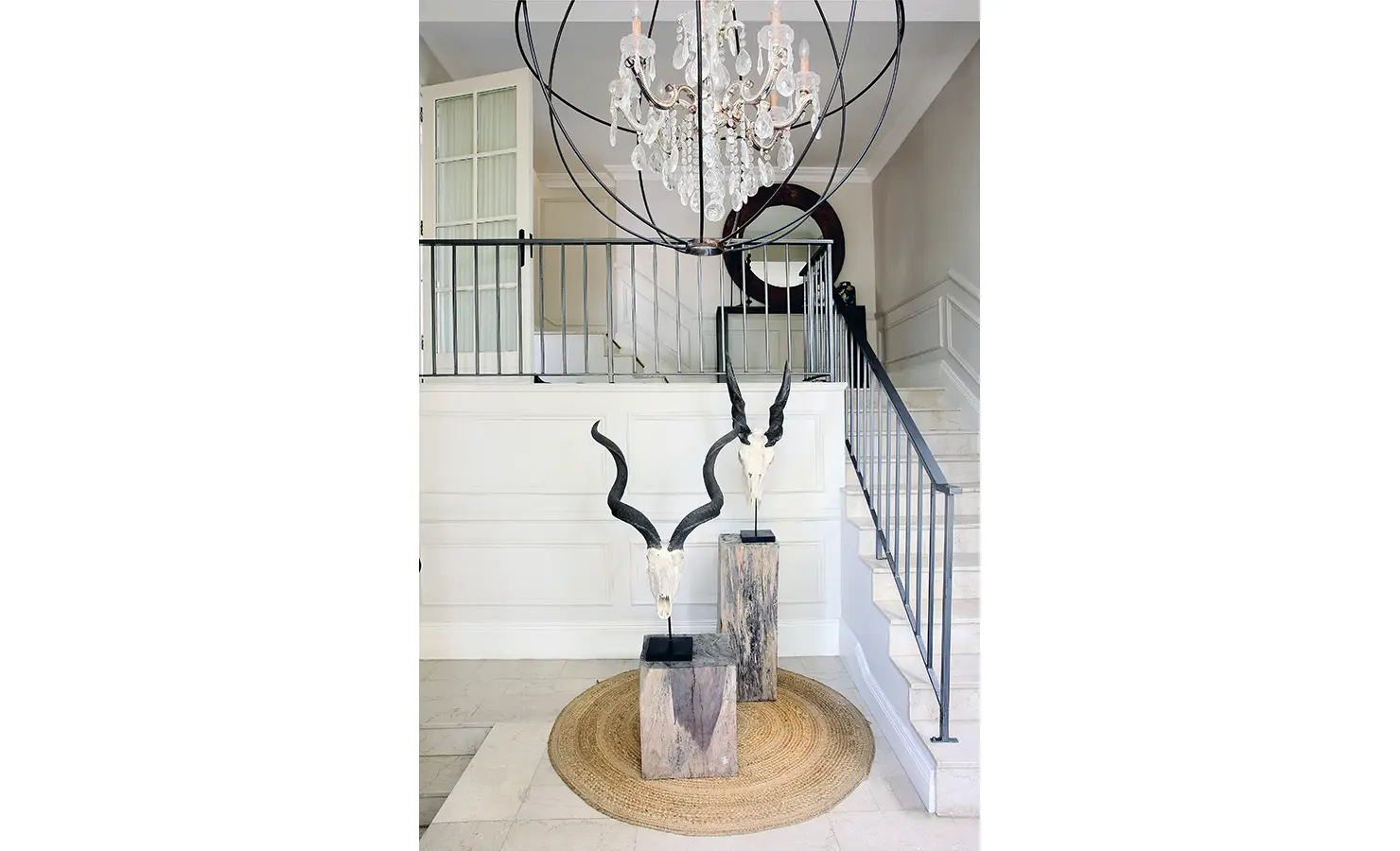 Perfect Hideaways: Double-volume entrance hall featuring kudu horn sculptures on rustic plinths, a woven rug, and a dramatic chandelier suspended above the staircase. Pepper Tree House, De Waterkant.