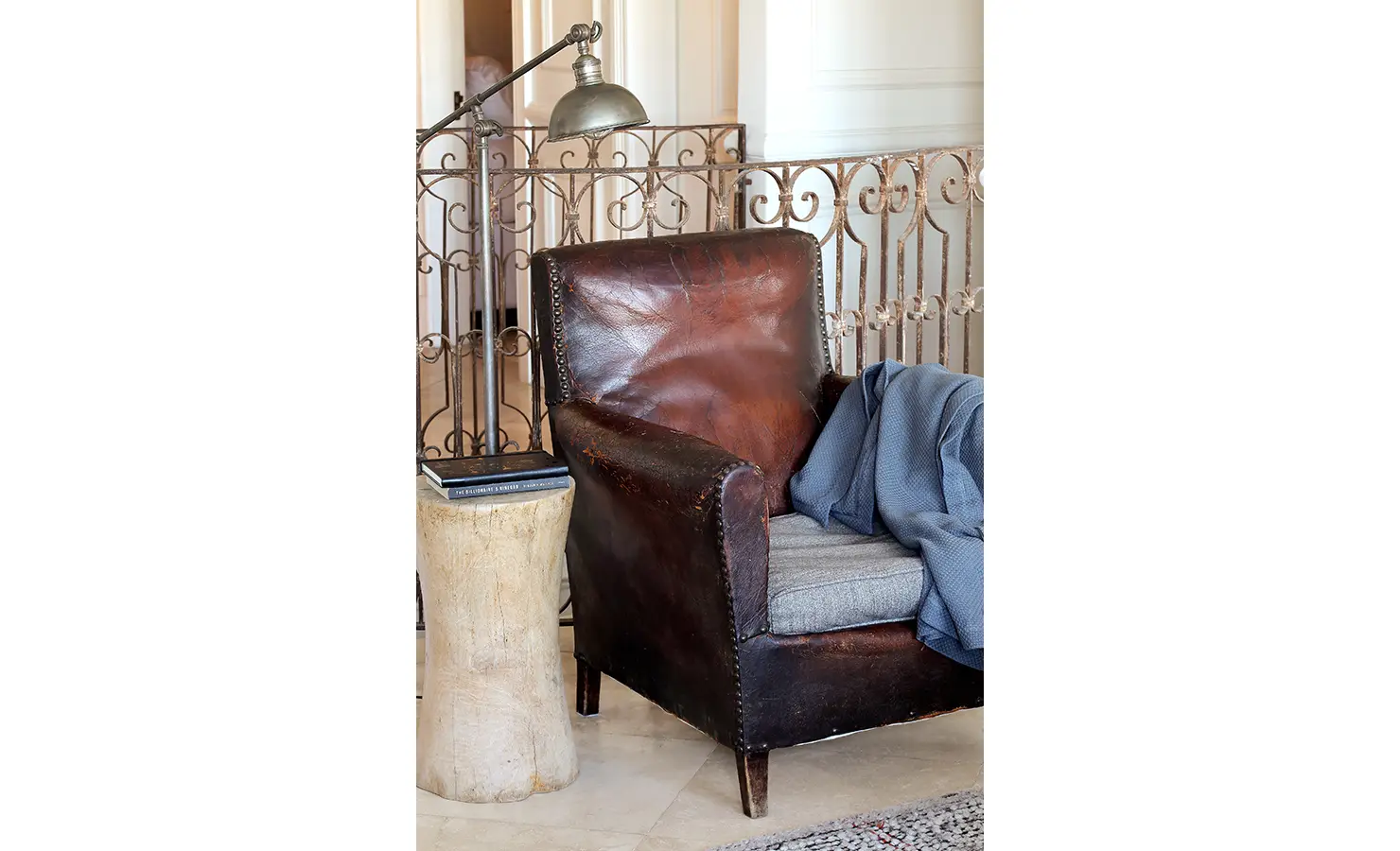 Perfect Hideaways: A worn leather armchair with grey cushion and blue throw stands beside a rustic stump side table and vintage industrial lamp, exuding timeless comfort. Pepper Tree House, De Waterkant.