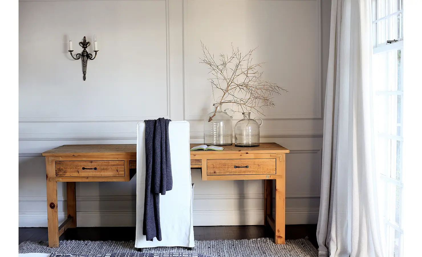 Perfect Hideaways: Minimalist writing desk in natural wood with glass demijohns and a bare branch arrangement, set against soft panelled walls and a window with flowing curtains. Pepper Tree House, De Waterkant.