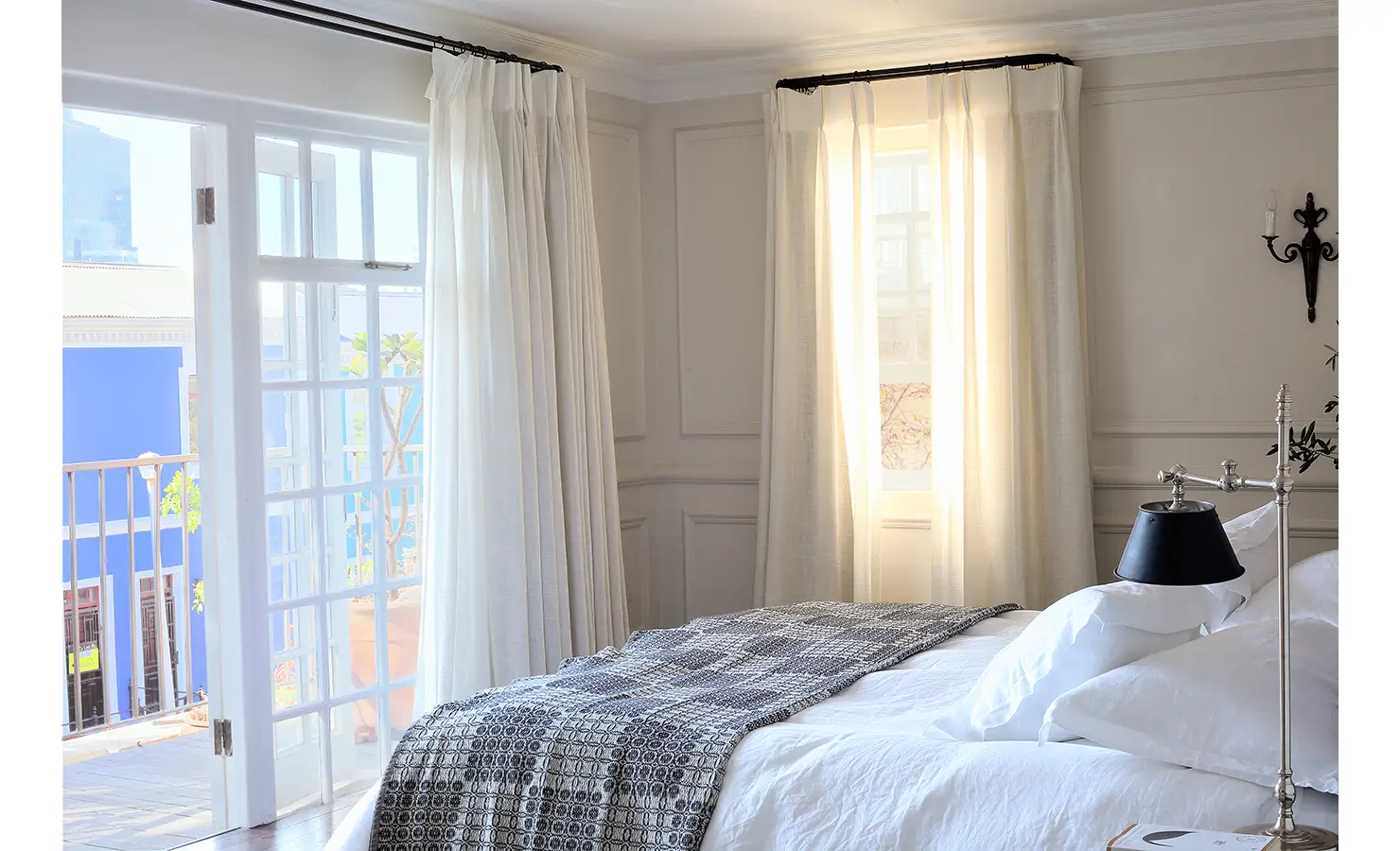 Perfect Hideaways: Serene bedroom bathed in morning light, with crisp white linens, iron-framed bed, and floor-to-ceiling curtains opening onto a balcony with vibrant Bo-Kaap views. Pepper Tree House, De Waterkant.