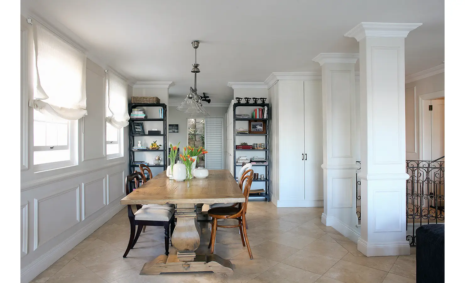 Perfect Hideaways: Open-plan dining space with a rustic wooden table, mismatched chairs, and shelves filled with books and curiosities, all softened by white Roman blinds. Pepper Tree House, De Waterkant.