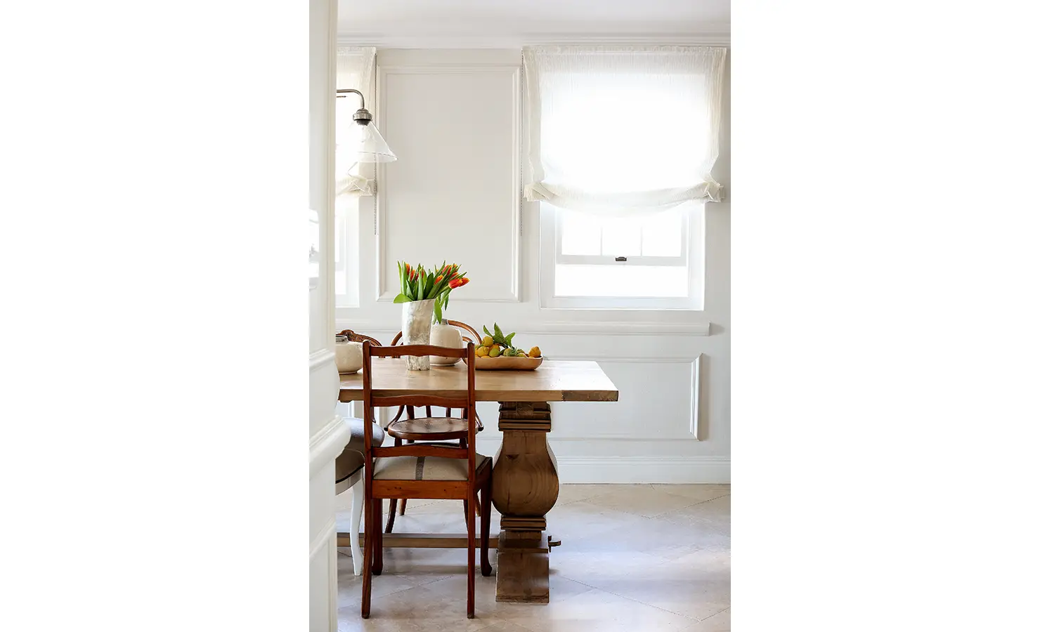 Perfect Hideaways: A sunlit dining nook with a rustic wooden table, vintage chairs, and tulips in a marble vase, framed by soft Roman blinds and classic wall panelling. Pepper Tree House, De Waterkant.
