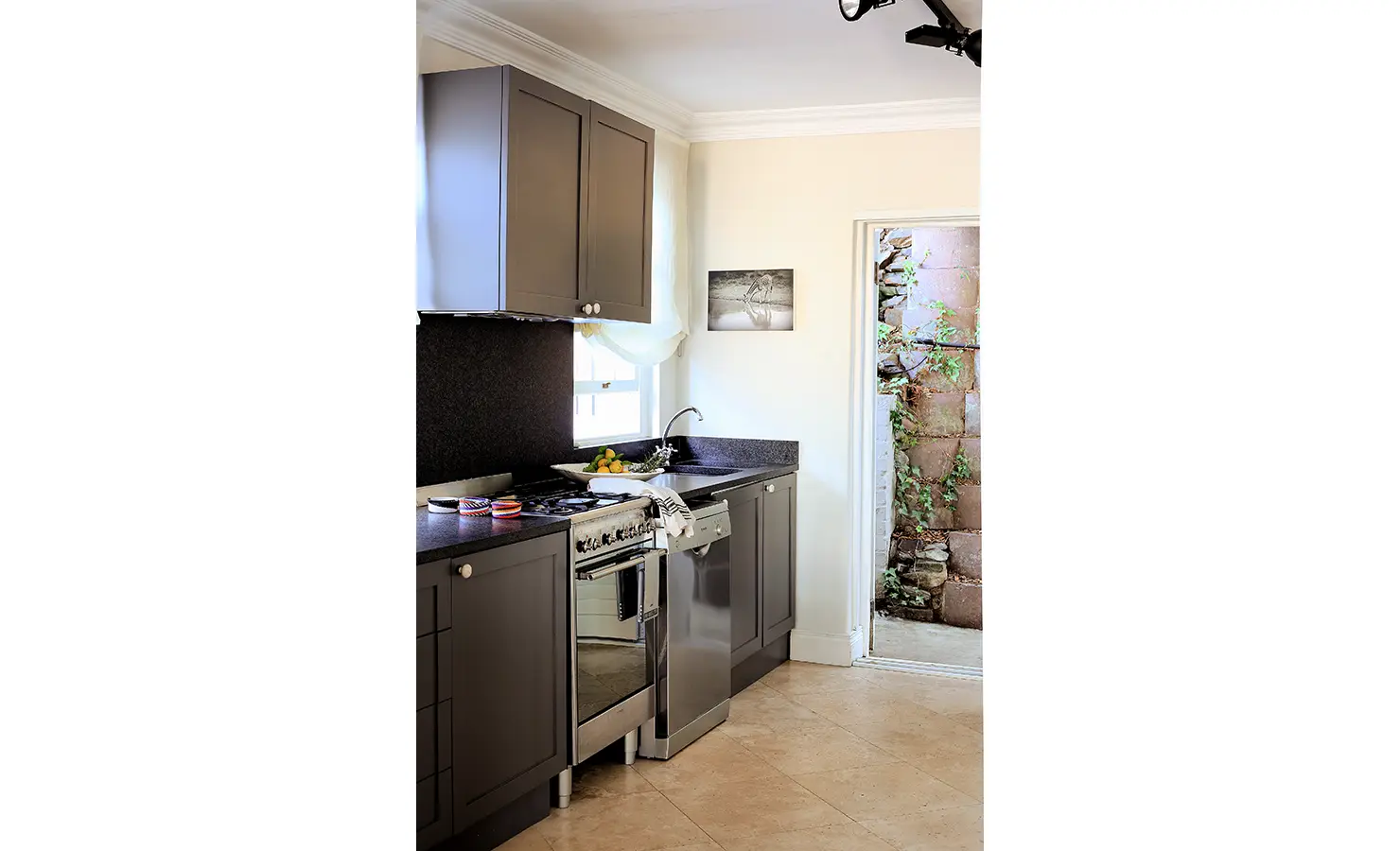 Perfect Hideaways: Compact kitchen with charcoal cabinetry, stainless steel appliances, and a window looking out to a vine-covered stone wall. Pepper Tree House, De Waterkant.