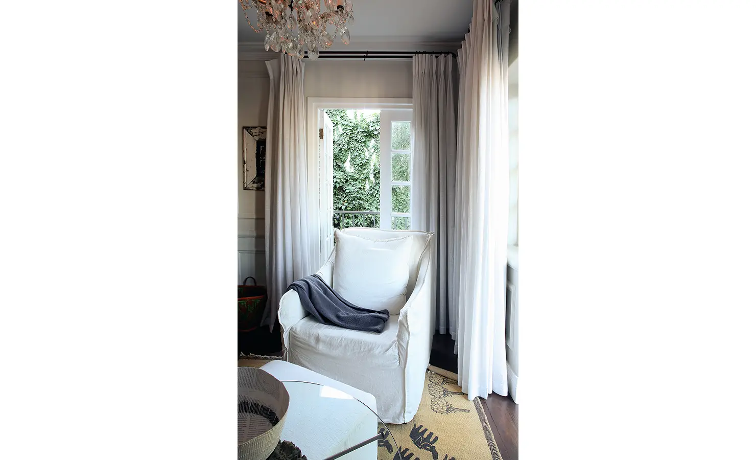 Perfect Hideaways: A cosy white armchair with a dark throw tucked into a corner framed by flowing curtains and French doors opening to lush greenery. Pepper Tree House, De Waterkant.