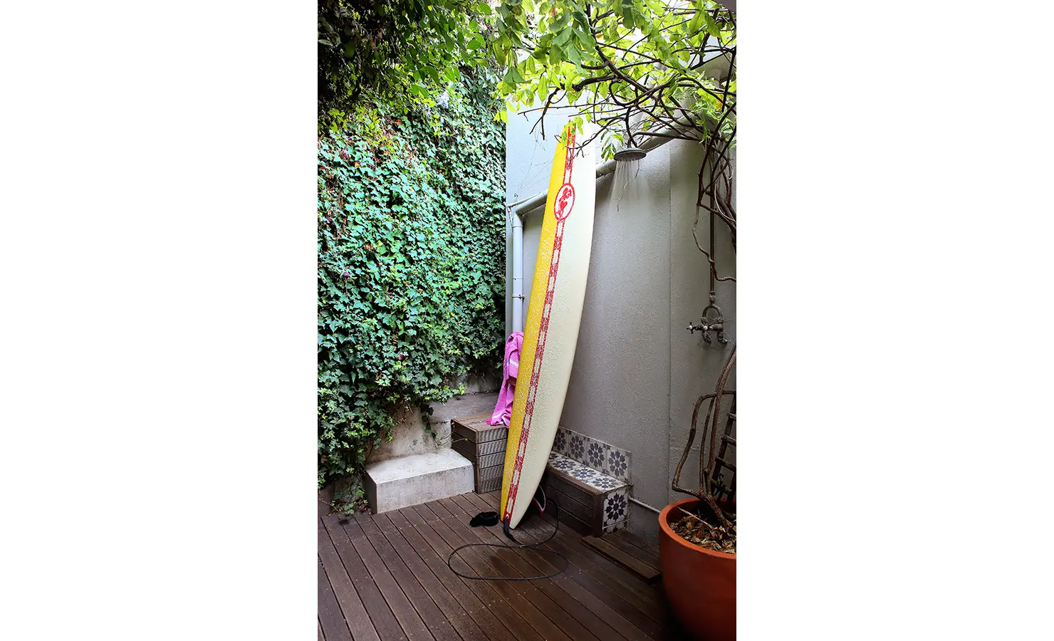 Perfect Hideaways: Ivy-clad outdoor shower area with wooden decking, a surfboard leaning casually against the wall, and climbing vines overhead. Pepper Tree House, De Waterkant.