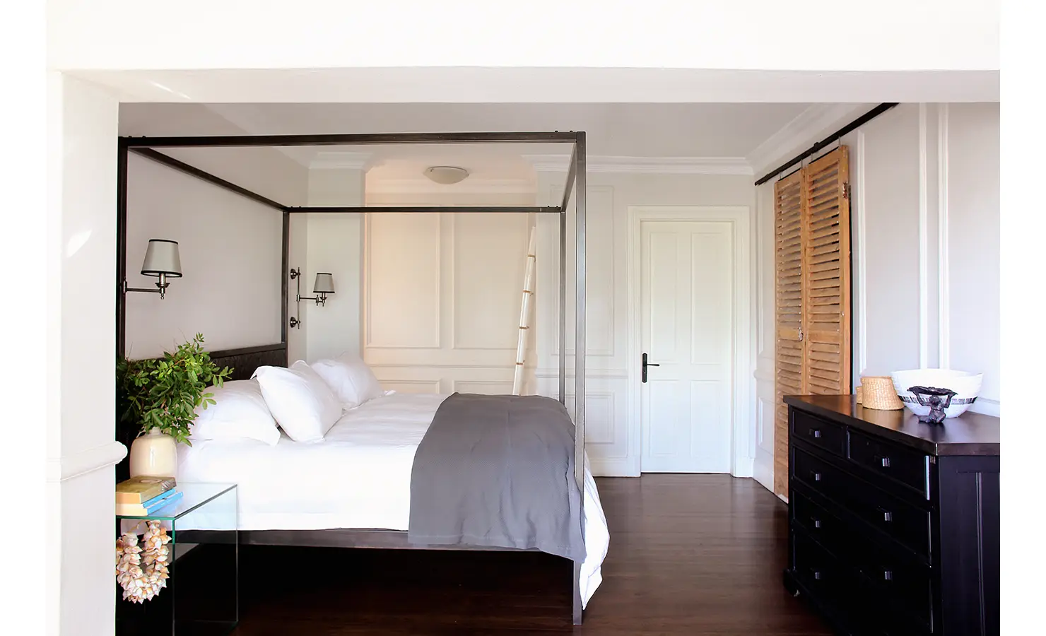 Perfect Hideaways: Bright bedroom with a sleek four-poster bed, minimalist décor, and natural accents like a woven shell garland and leafy plant. Pepper Tree House, De Waterkant.