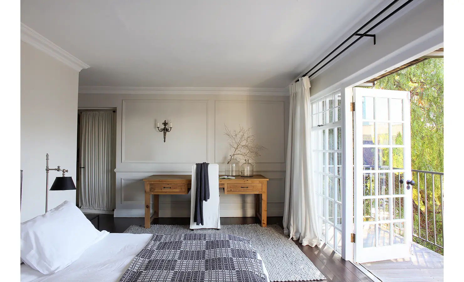 Perfect Hideaways: Airy bedroom corner with French doors opening onto a balcony, a natural wood desk, and soft layers of white and grey creating a tranquil retreat. Pepper Tree House, De Waterkant.