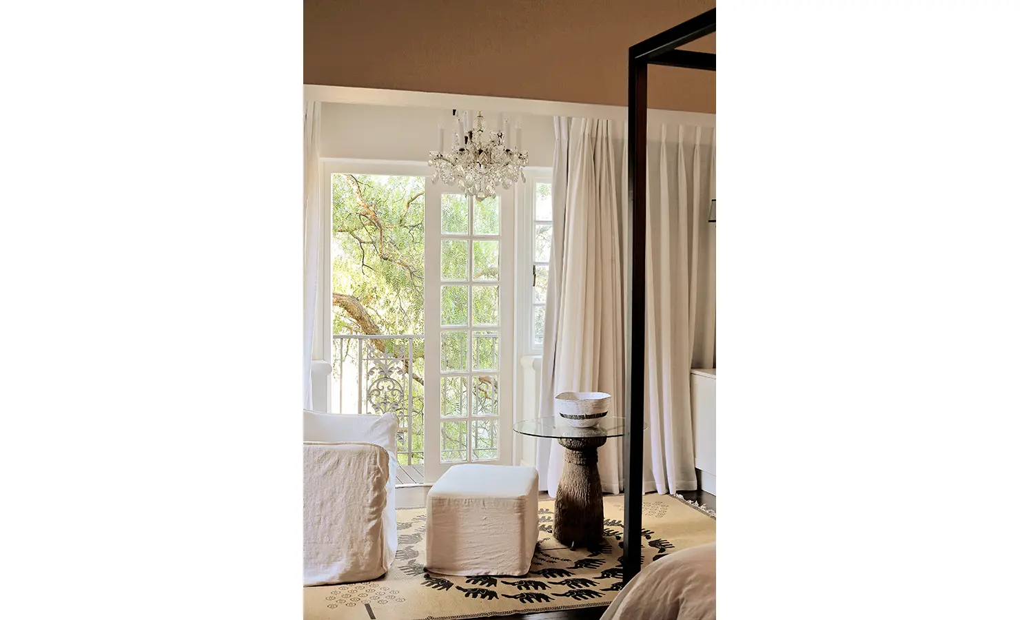 Perfect Hideaways: Serene bedroom corner with draped curtains, a crystal chandelier, and slipcovered seating, opening onto a leafy balcony dappled with soft natural light. Pepper Tree House, De Waterkant.