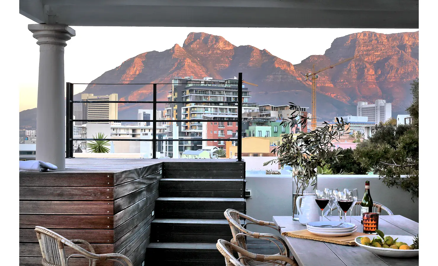 Perfect Hideaways: Outdoor dining terrace with a rustic wooden table set for wine and citrus, framed by urban greenery and views of Table Mountain glowing in the evening light. Pepper Tree House, De Waterkant.