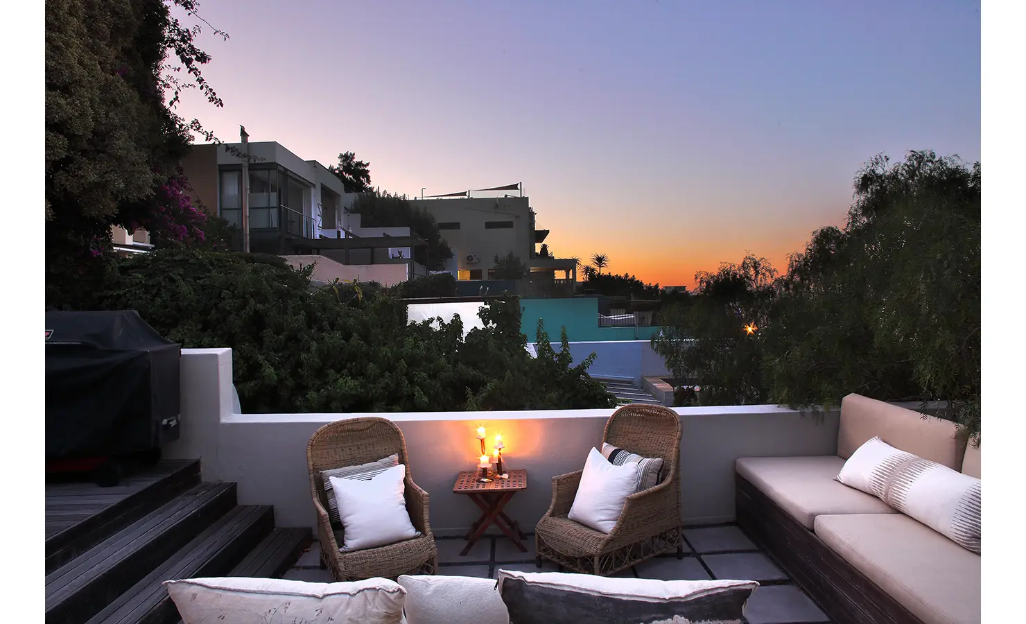 Perfect Hideaways: Intimate rooftop lounge at dusk with woven armchairs, plush cushions, and candlelight, set against a tranquil sunset and modern hillside homes. Pepper Tree House, De Waterkant.
