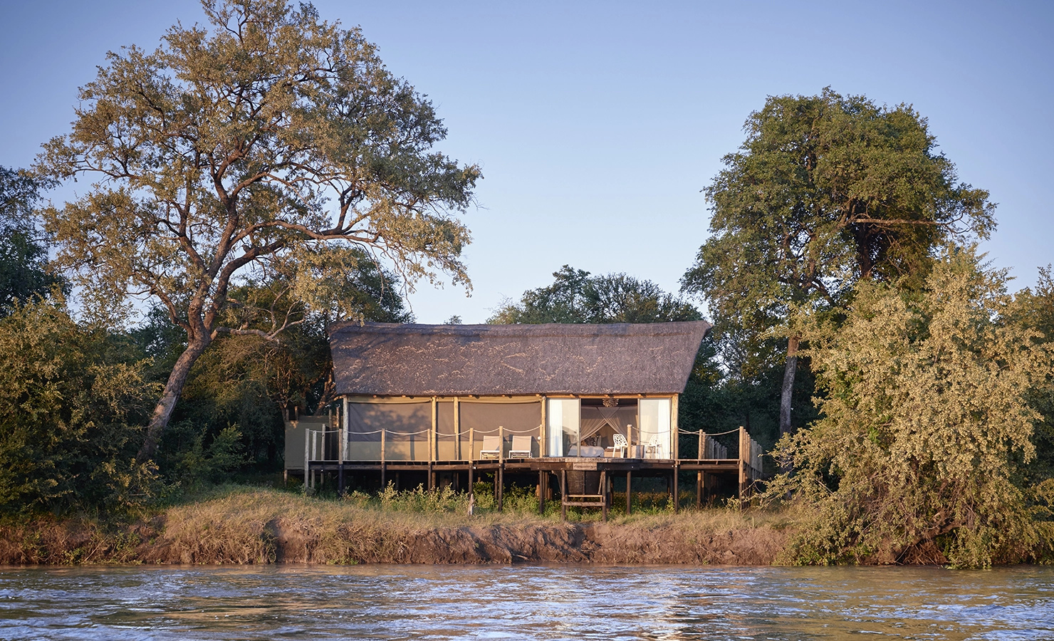 Perfect Hideaways: A luxury safari tent with a thatched roof stands elevated on a wooden deck overlooking the Zambezi River, surrounded by lush trees and bathed in golden sunlight. White outdoor chairs invite relaxation on the veranda, blending seamlessly with the natural beauty of the wilderness. Victoria Falls River Lodge, Zambezi National Park.