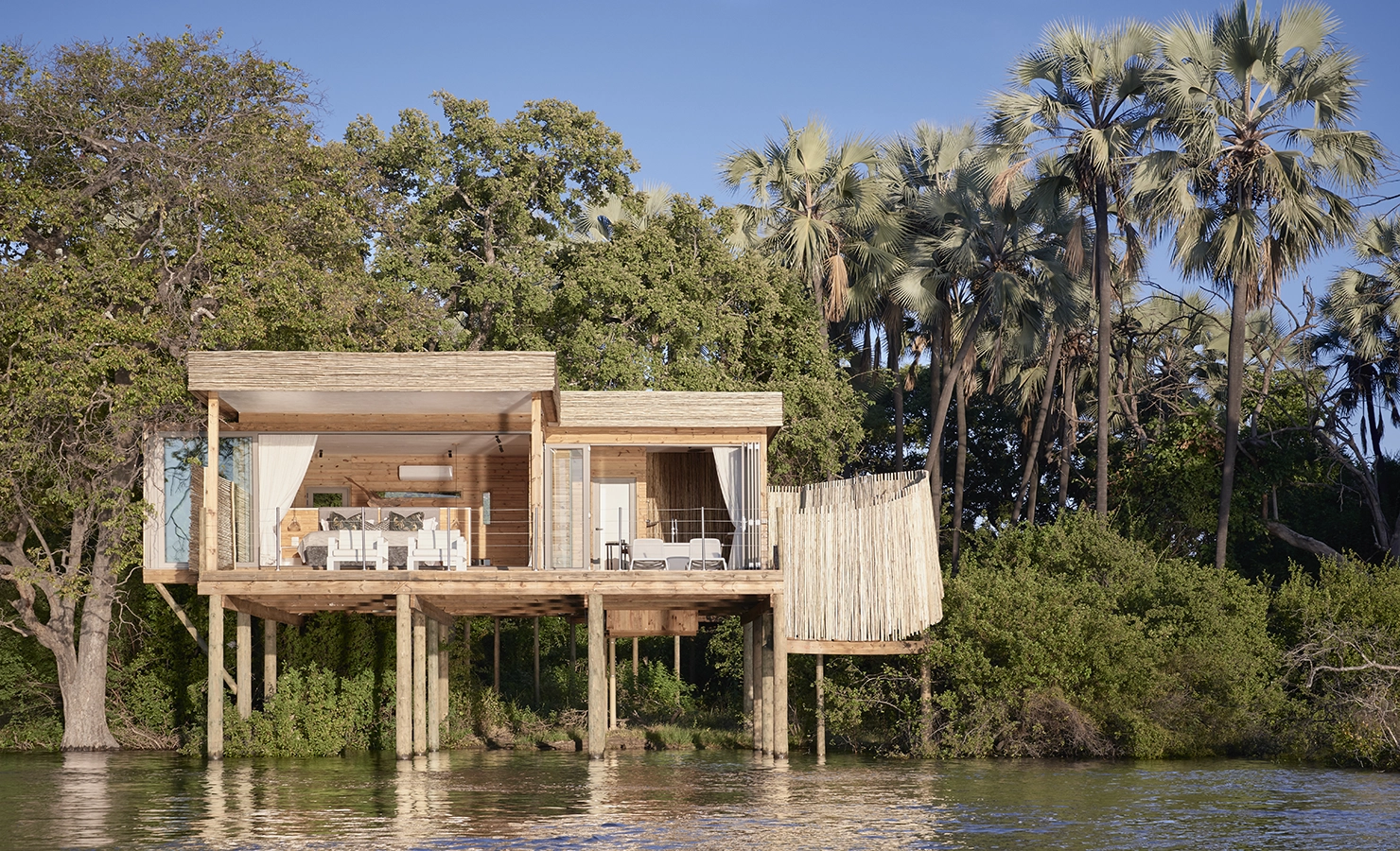 Perfect Hideaways: A luxurious wooden safari lodge elevated on stilts above the Zambezi River, blending seamlessly with the lush riverine forest and palm trees, offering an open-plan design with elegant furnishings and a private deck. Victoria Falls Island Lodge, Zambezi National Park.