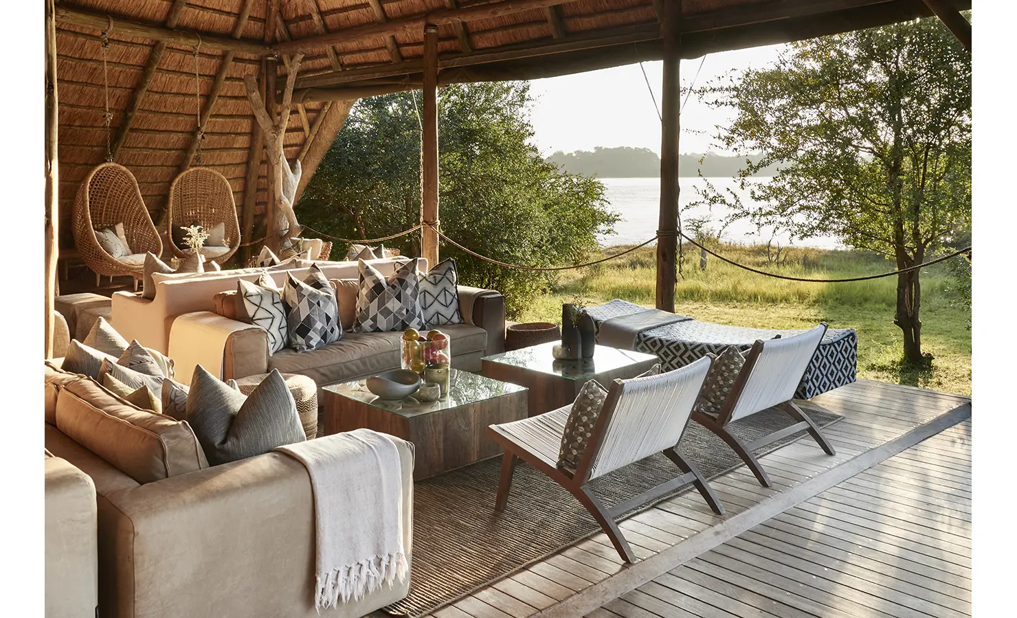 Perfect Hideaways: A beautifully designed open-air lounge under a thatched roof, with plush sofas, patterned cushions, and hanging rattan chairs, overlooking the tranquil waters of the Zambezi River. Victoria Falls River Lodge, Zambezi National Park.