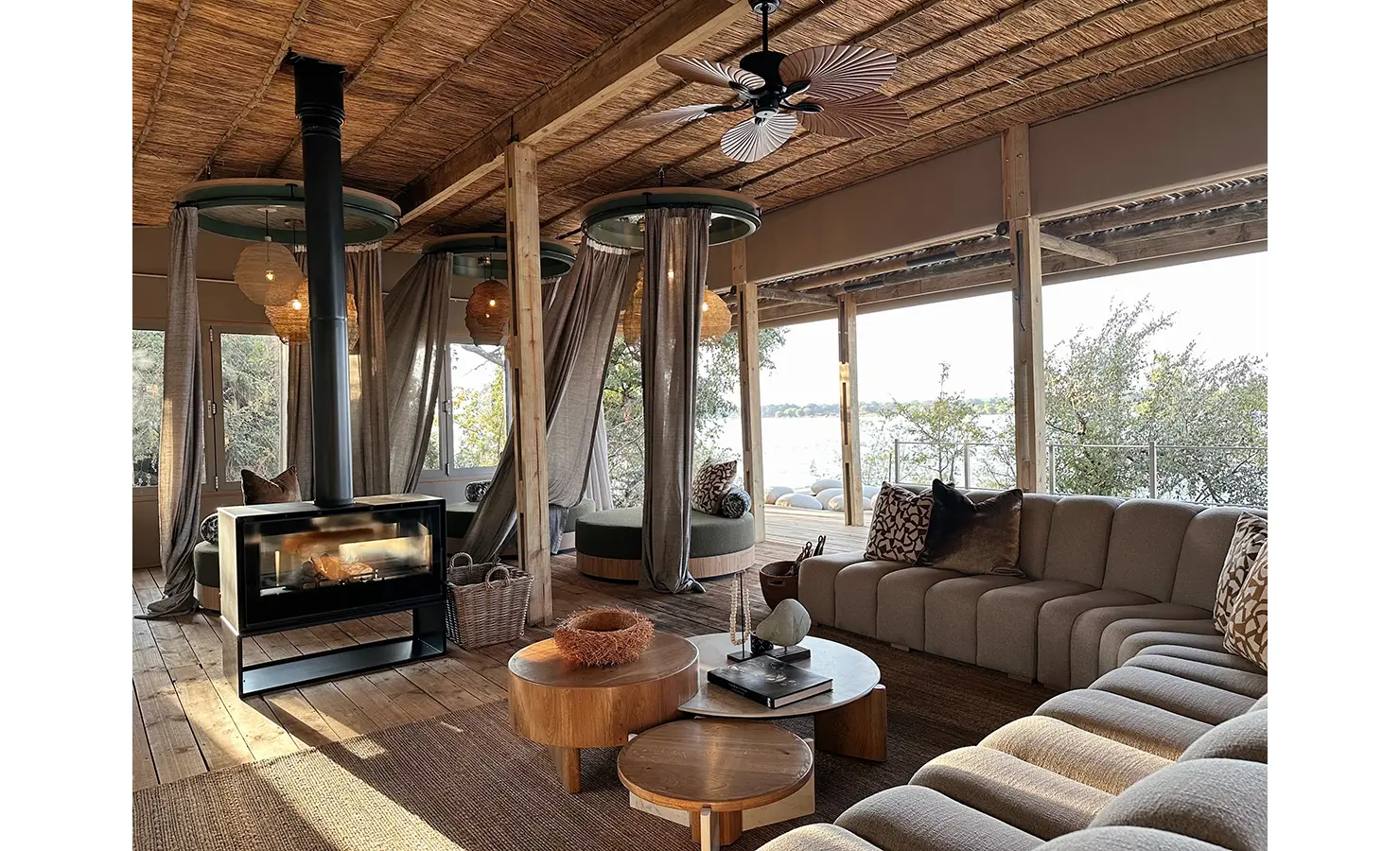 Perfect Hideaways: A stylishly decorated indoor lounge with a woven ceiling, an elegant fireplace, and soft curved sofas, creating a warm and inviting space with panoramic views of the river beyond. Victoria Falls River Lodge, Zambezi National Park.