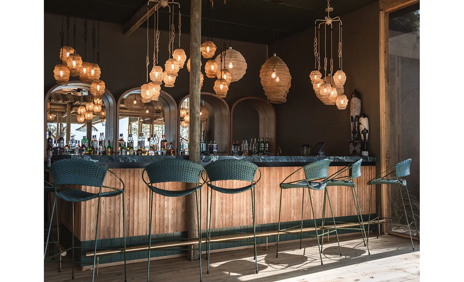 Perfect Hideaways: A chic bar area with a natural wood counter, green barstools, and an array of unique pendant lights, offering an elegant setting for evening drinks with river views. Victoria Falls River Lodge, Zambezi National Park.