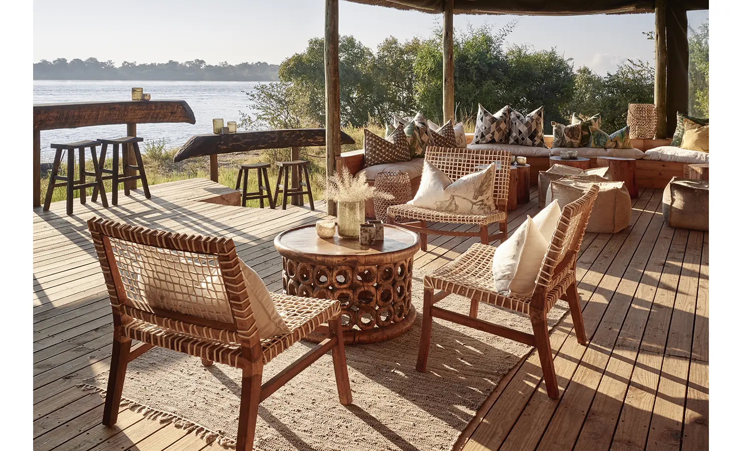 Perfect Hideaways: An inviting open-air deck furnished with woven chairs and wooden tables, offering a relaxed riverside retreat with uninterrupted views of the flowing Zambezi River. Victoria Falls River Lodge, Zambezi National Park.