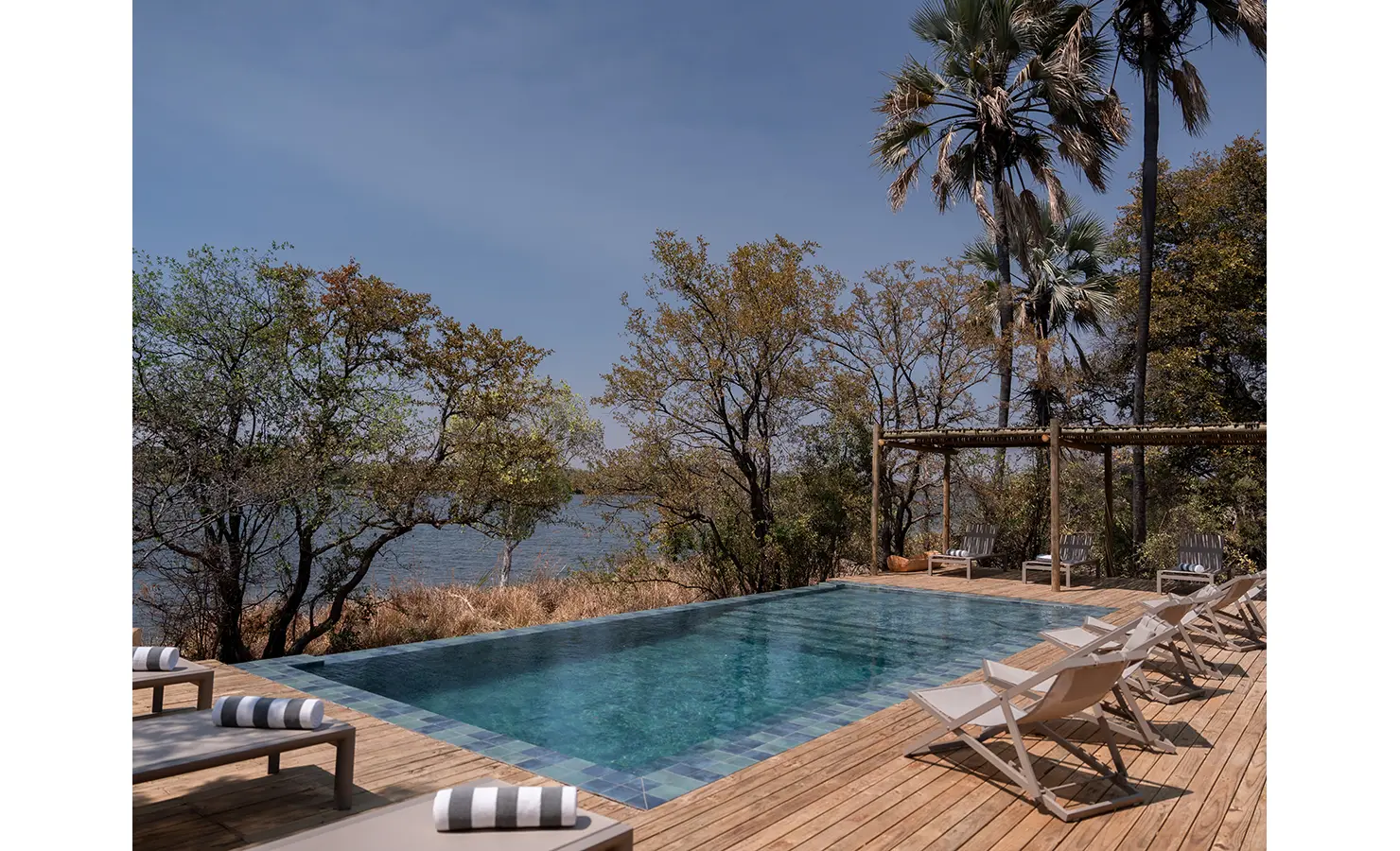 Perfect Hideaways: A serene poolside setting with a wooden deck lined with sun loungers, surrounded by indigenous trees, and overlooking the calm waters of the Zambezi River. Victoria Falls River Lodge, Zambezi National Park.