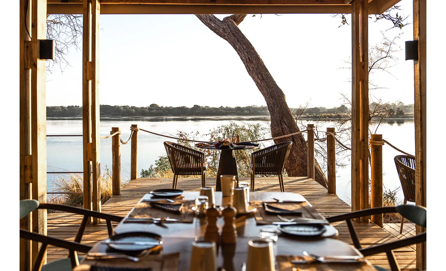 Perfect Hideaways: A beautifully set dining area on a wooden deck, with a table adorned with tableware and a private riverside seating arrangement, creating a magical setting for alfresco meals. Victoria Falls River Lodge, Zambezi National Park.