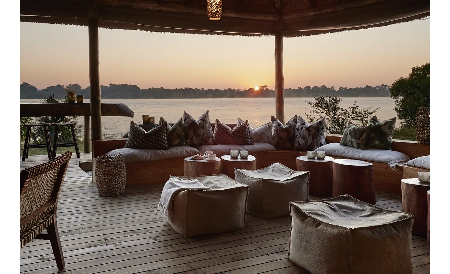 Perfect Hideaways: A cosy outdoor lounge at sunset, featuring a built-in seating area with patterned cushions and woven poufs, creating a relaxed atmosphere with breathtaking river views. Victoria Falls River Lodge, Zambezi National Park.