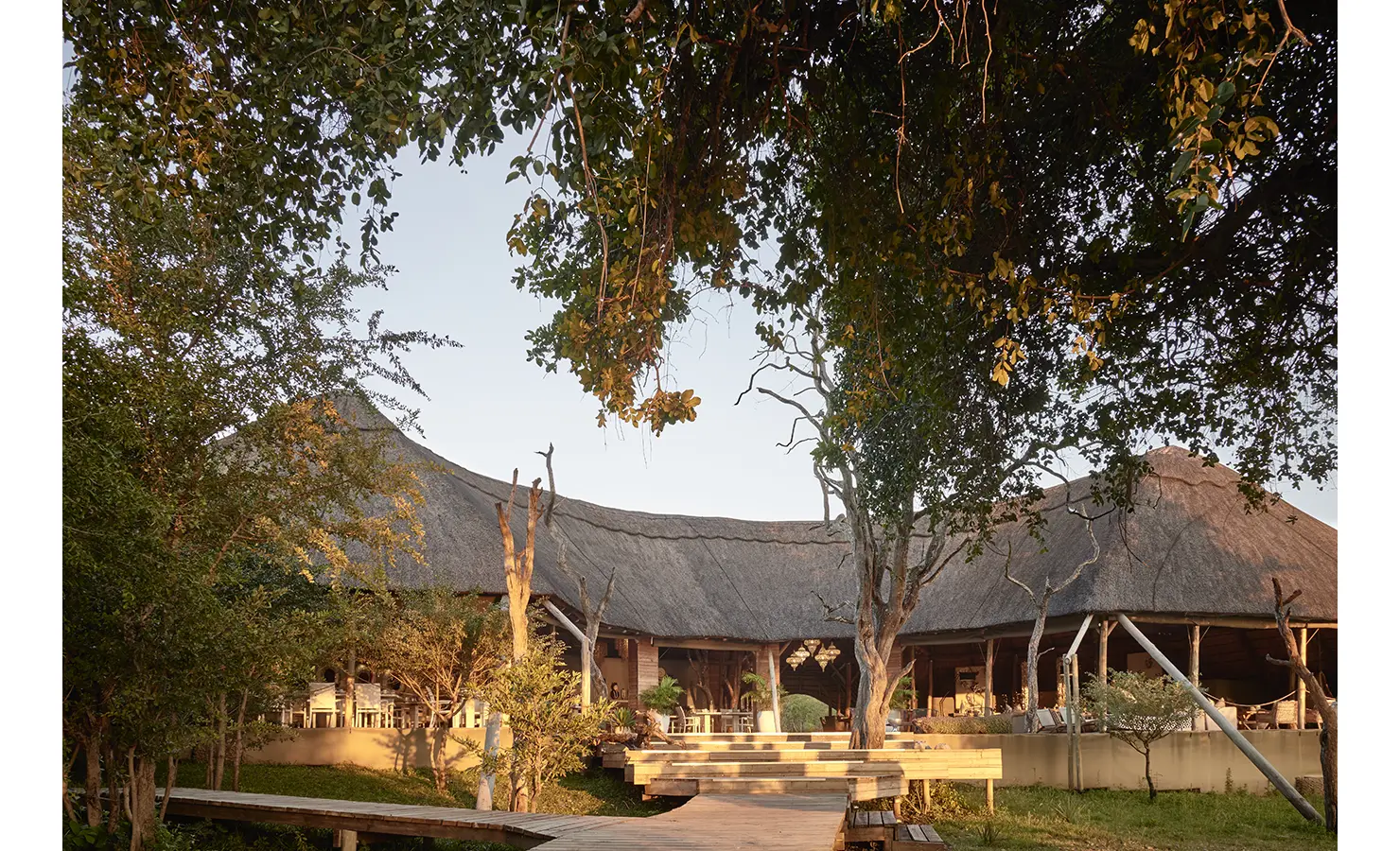 Perfect Hideaways: A stunning thatched-roof main lodge set amongst lush greenery, with a welcoming wooden walkway leading to the open-plan living and dining areas. Victoria Falls River Lodge, Zambezi National Park.