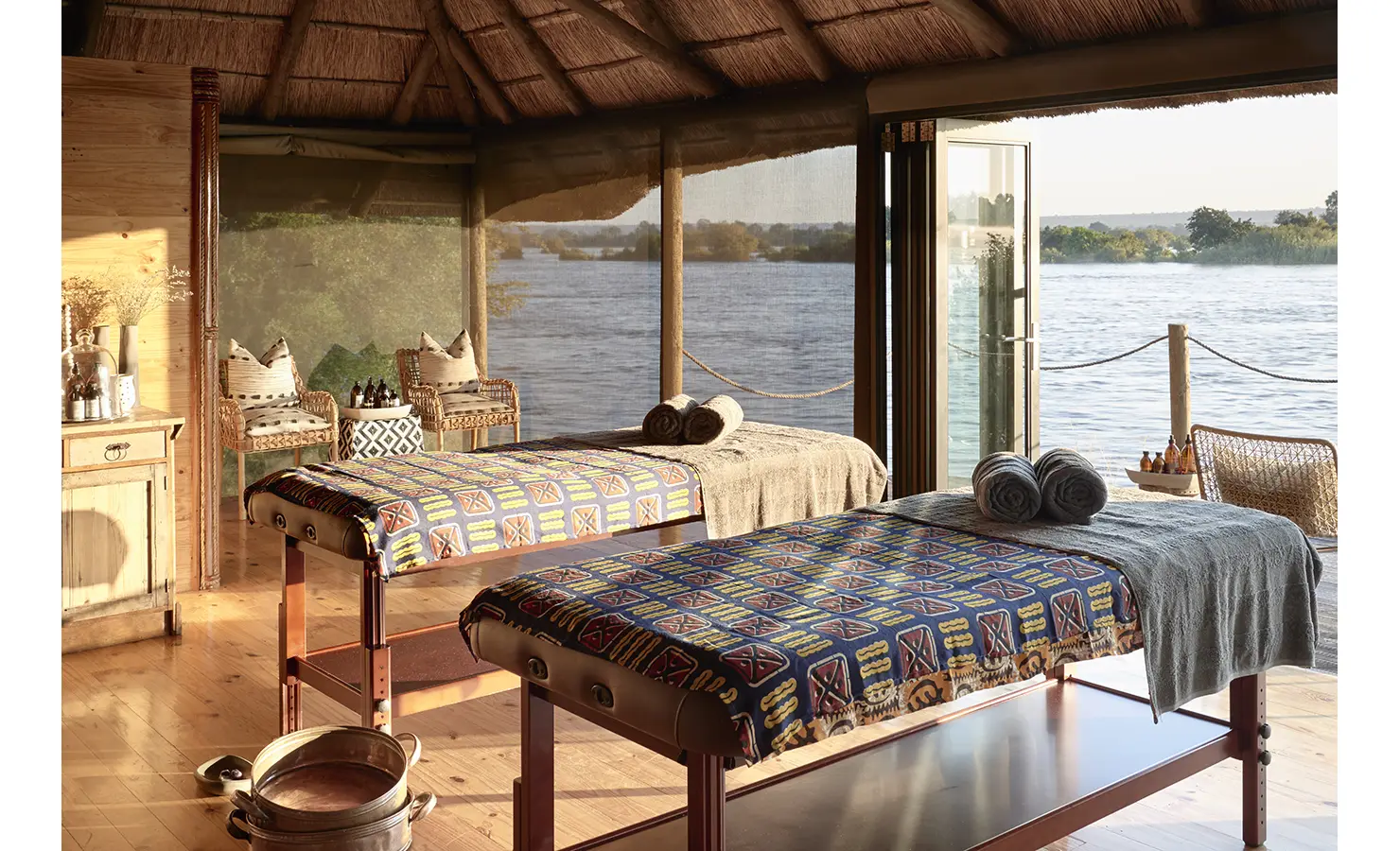 Perfect Hideaways: A tranquil spa treatment room with wooden massage tables, adorned with vibrant textiles, positioned beside large windows that open to sweeping river views. Victoria Falls River Lodge, Zambezi National Park.