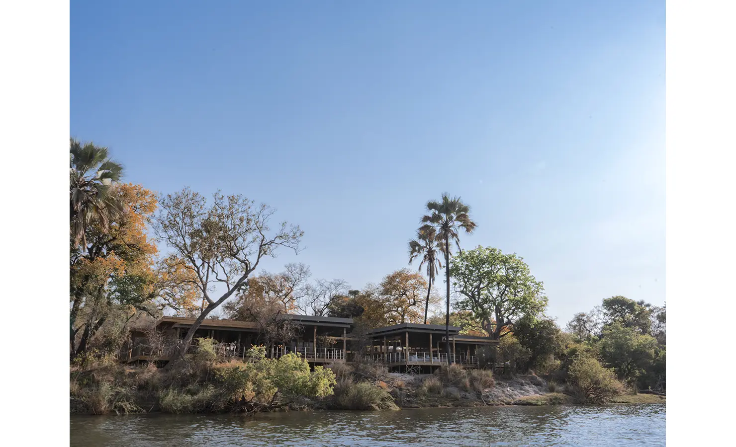 Perfect Hideaways: A stunning riverside lodge, nestled among lush trees and towering palms, elevated above the Zambezi River with expansive decks that blend seamlessly into the natural surroundings. Victoria Falls River Lodge, Zambezi National Park.
