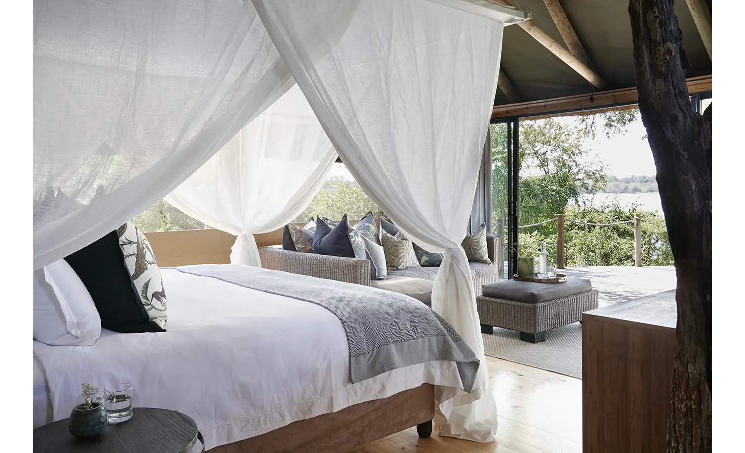 Perfect Hideaways: A luxurious safari bedroom with a four-poster bed draped in white linens, flowing sheer curtains, and an inviting lounge area leading onto a private deck with views of the Zambezi River. Victoria Falls River Lodge, Zambezi National Park.