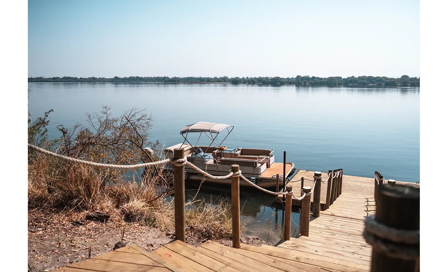 Perfect Hideaways: A tranquil riverside dock with a moored pontoon boat, accessed by a wooden walkway, offering an idyllic setting for leisurely river cruises. Victoria Falls River Lodge, Zambezi National Park.