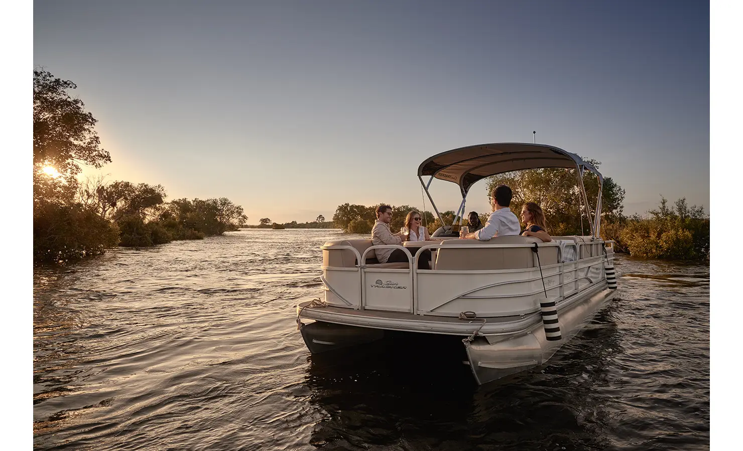 Perfect Hideaways: A group of guests enjoying sundowner drinks aboard a luxury pontoon boat, cruising the Zambezi River at golden hour as the sun sets behind the trees. Victoria Falls River Lodge, Zambezi National Park.