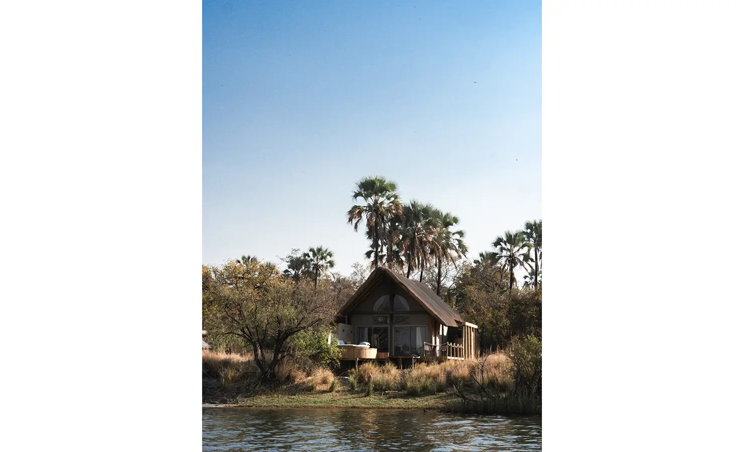 Perfect Hideaways: A charming thatched-roof safari villa, tucked among palm trees on the riverbank, offering seclusion and serenity amidst the African wilderness. Victoria Falls River Lodge, Zambezi National Park.