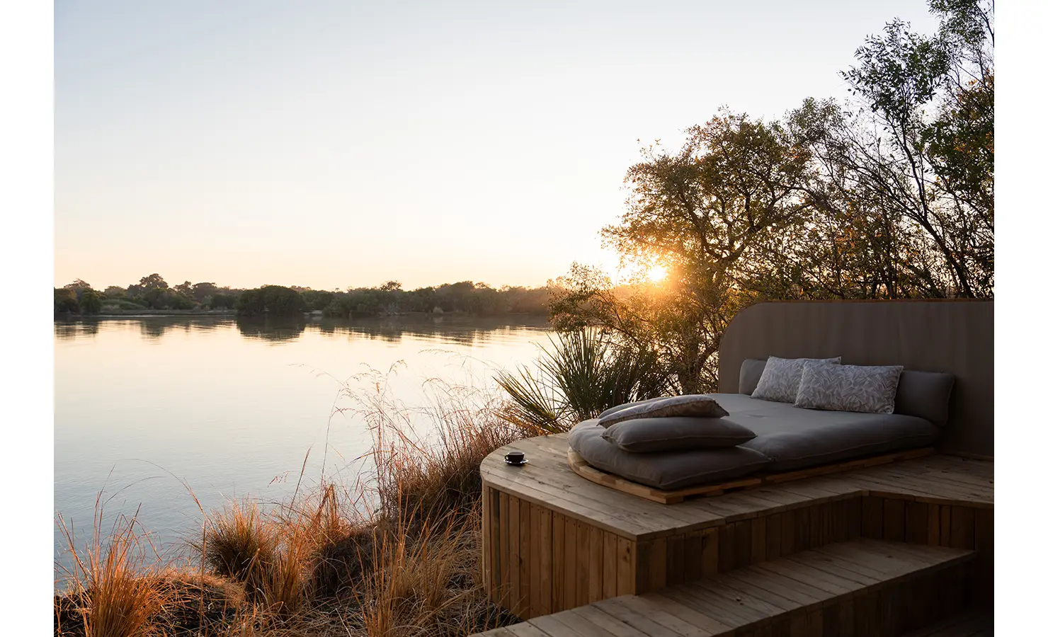 Perfect Hideaways: A dreamy riverside relaxation deck with a luxurious daybed, set against a breathtaking sunset over the Zambezi River, offering an intimate retreat into nature. Victoria Falls River Lodge, Zambezi National Park.