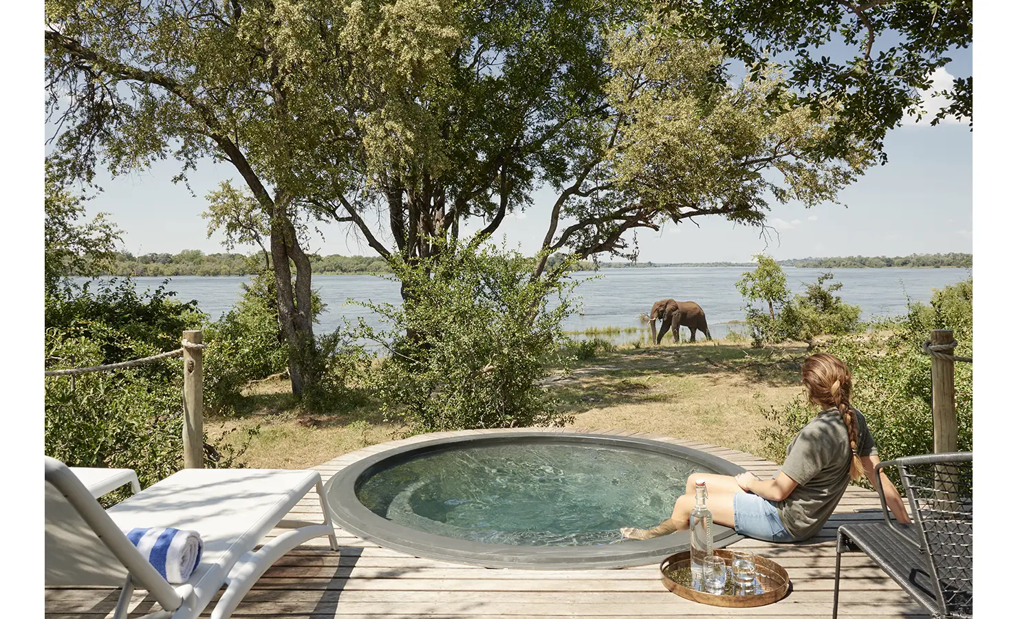 Perfect Hideaways: A peaceful private plunge pool on a wooden deck, where a guest relaxes while watching an elephant drink from the river just beyond the trees. Victoria Falls River Lodge, Zambezi National Park.