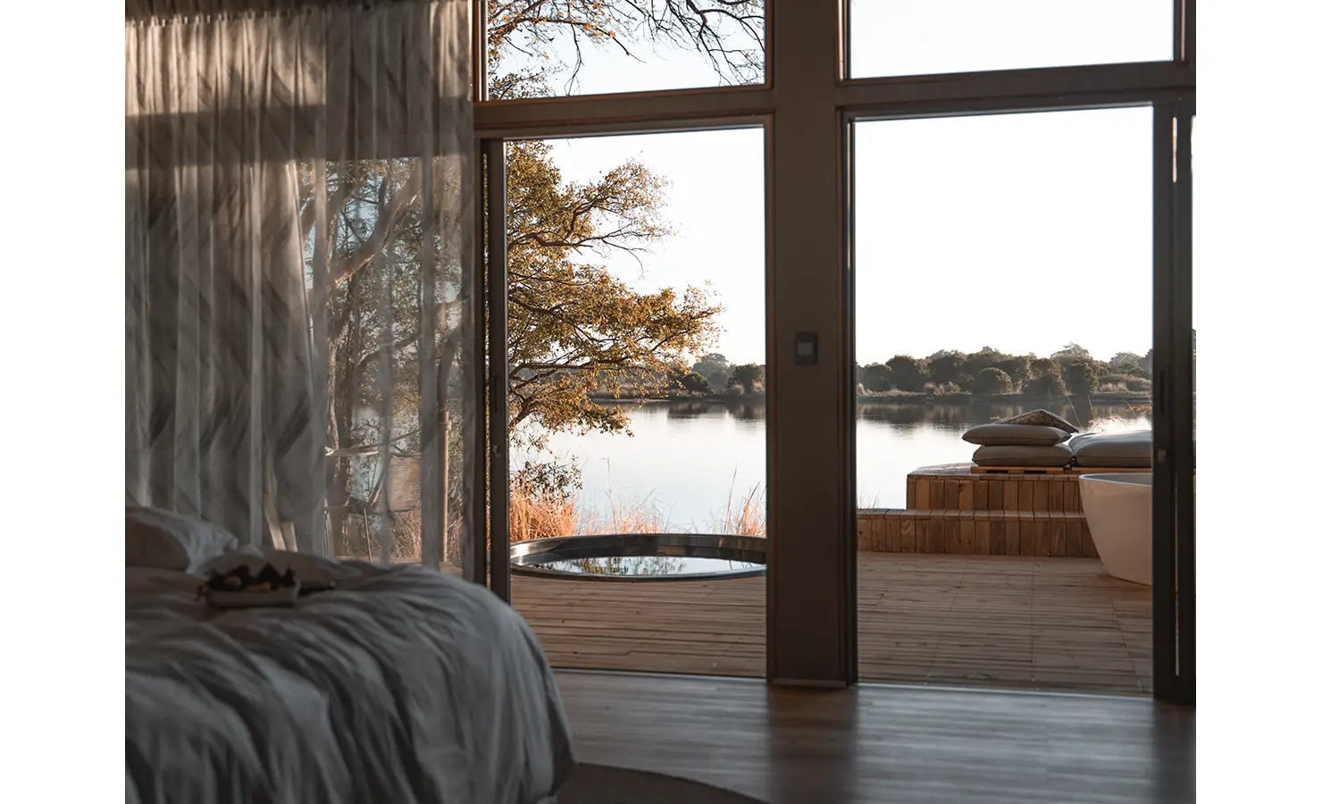 Perfect Hideaways: A serene bedroom view with sheer curtains swaying in the soft morning light, revealing a peaceful riverside deck with a plunge pool and a daybed overlooking the calm waters. Victoria Falls River Lodge, Zambezi National Park.