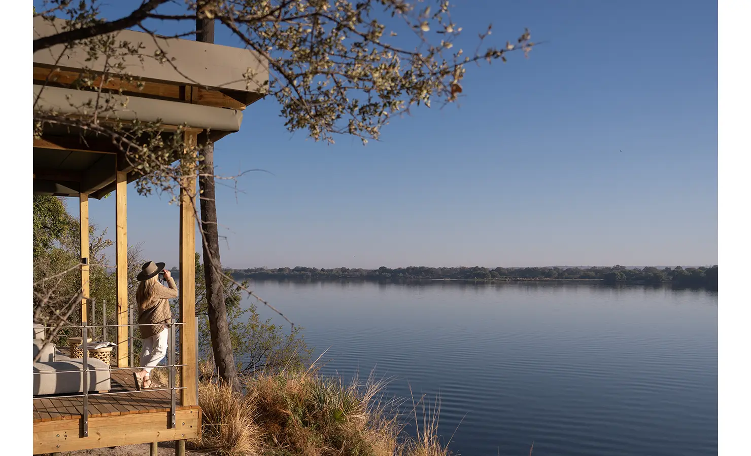 Perfect Hideaways: A guest standing on a private safari lodge deck, gazing out over the vast expanse of the Zambezi River, framed by golden grasses and lush trees. Victoria Falls River Lodge, Zambezi National Park.