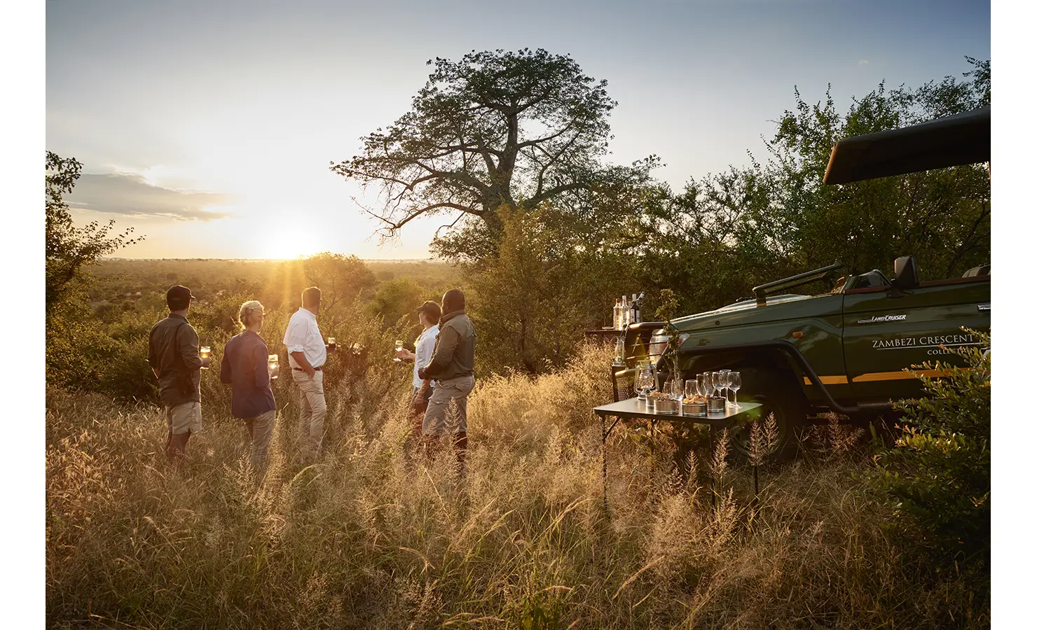Perfect Hideaways: A magical safari sundowner experience, with guests gathered around a drinks table set beside an open safari vehicle, enjoying cocktails as the sun sets over the vast African landscape. Victoria Falls River Lodge, Zambezi National Park.