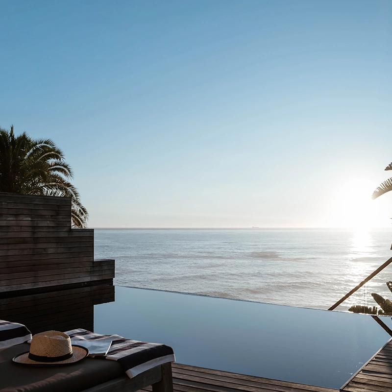 Perfect Hideaways oceanfront view at sunset featuring an infinity pool overlooking calm seas, with a woman in a flowing white dress walking along a wooden deck surrounded by palm trees and coastal tranquility, Bantry Bay, Arcadia.