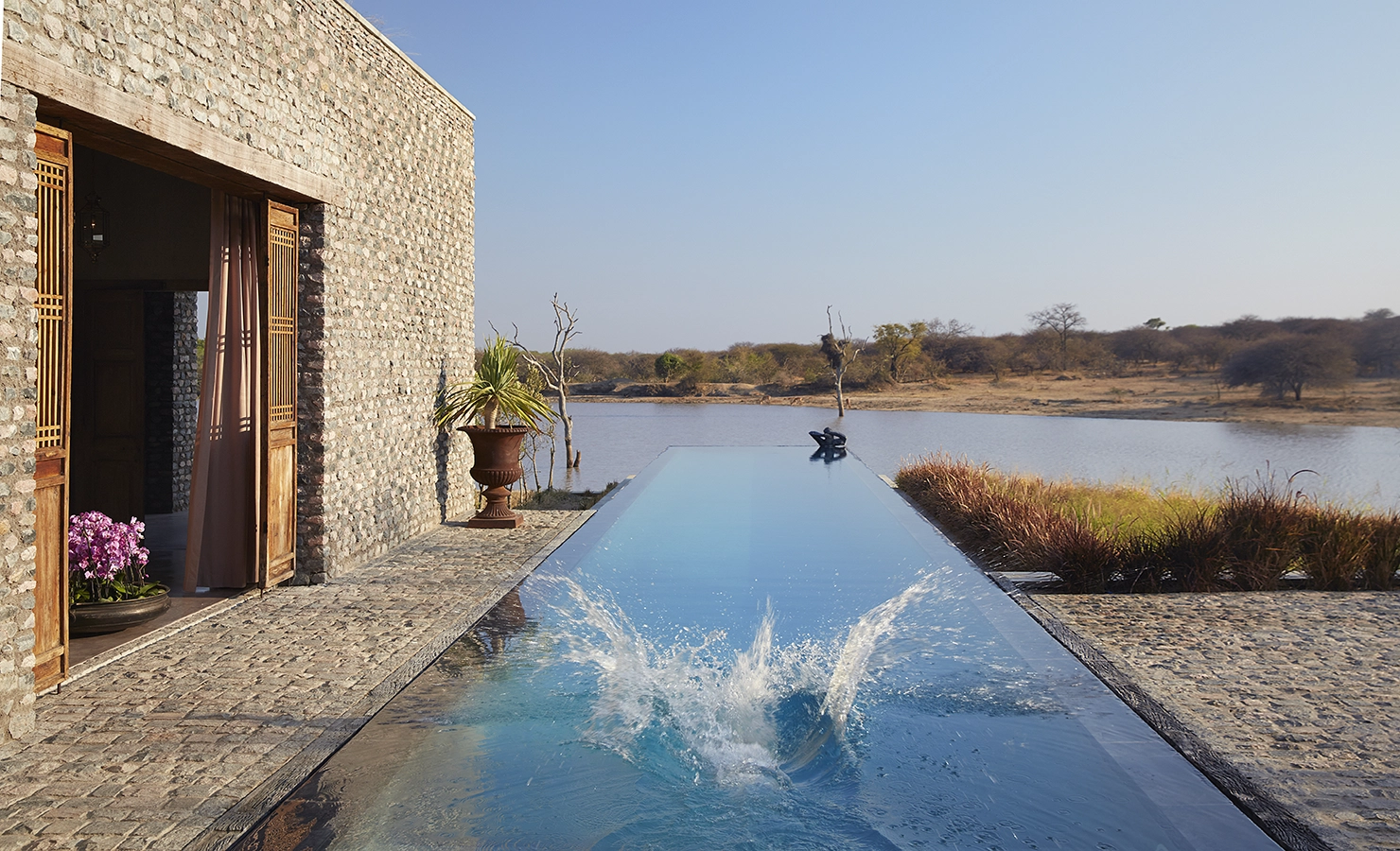 Perfect Hideaways: An inviting infinity pool extending towards the river, framed by a stone patio and lush greenery, with a splash in the water adding a lively touch. Kubili House, Thornybush Game Reserve.