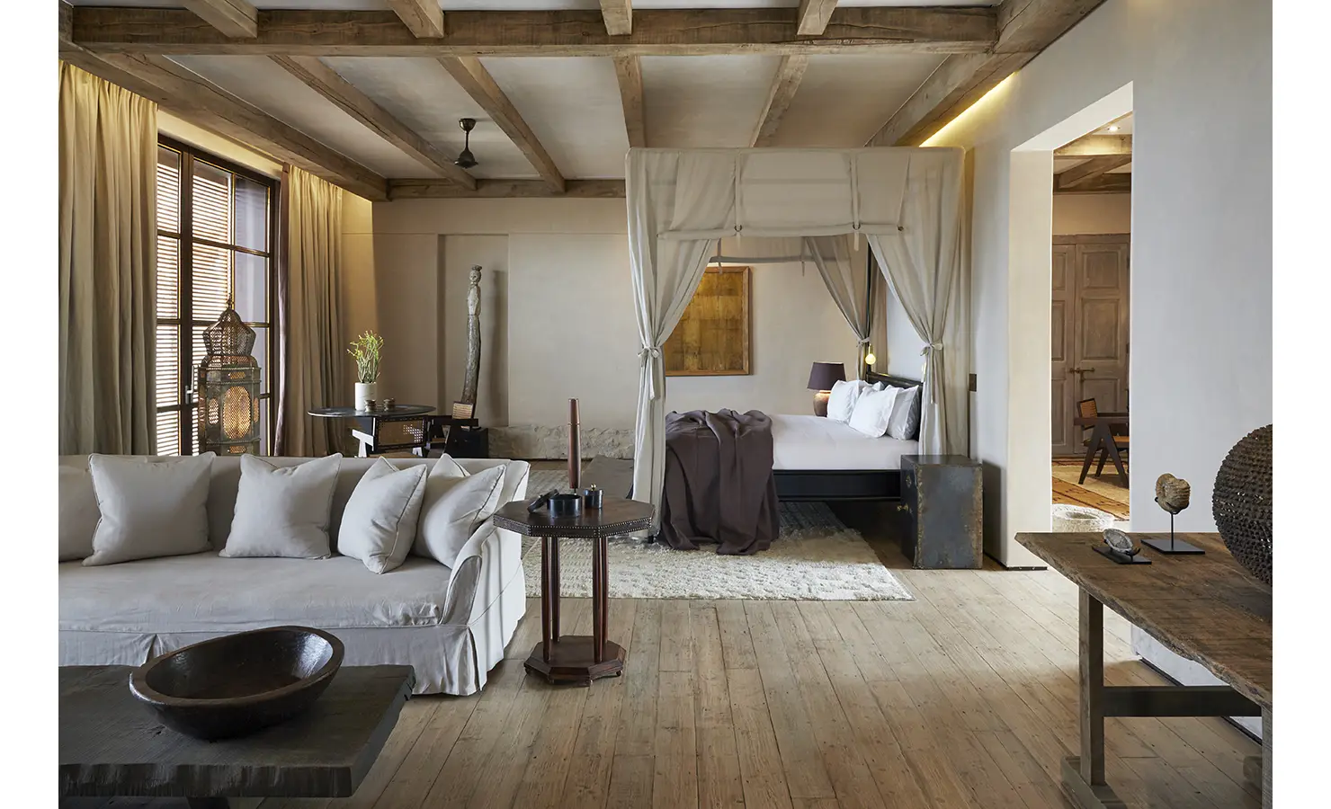 Perfect Hideaways: A luxurious bedroom with a four-poster bed draped in soft white curtains, a plush sofa, and rustic wooden beams on the ceiling. Warm lighting and natural textures create a cosy, elegant atmosphere. Kubili House, Thornybush Game Reserve.